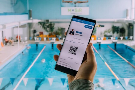 Dłoń trzymająca smartfon z&nbsp;aplikacją karty Rybka.W tle basen